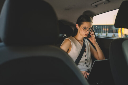 Businesswoman using a smartphone while working with a laptop in the back seat of a car