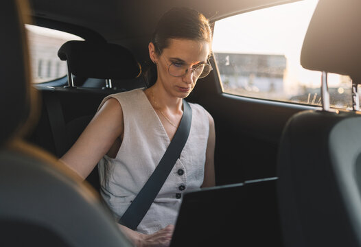 Businesswoman using a laptop while sitting with seat belt fastened in the back seat of a car 