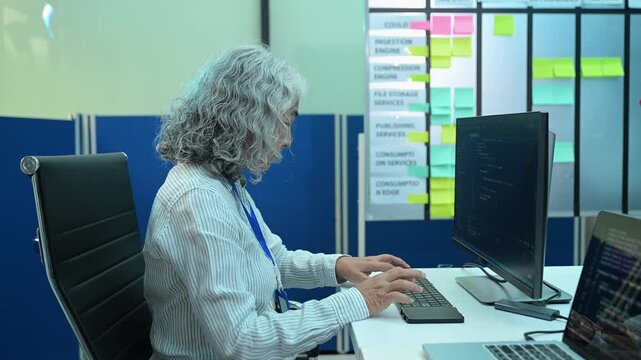 Senior Asian female software engineer coding on a desktop computer while a team member manages project tasks on an agile scrum board