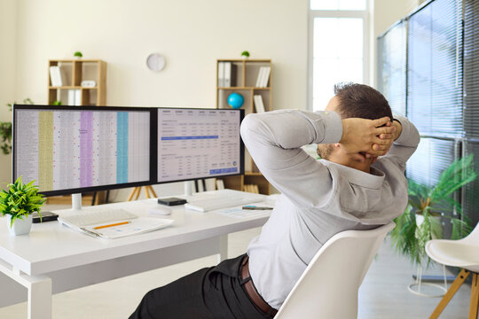 Employee analyst relax at office desk with spreadsheets. A man leans back at dual computer monitors after data analysis, taking a refreshing break. Clear concept of workplace success today.