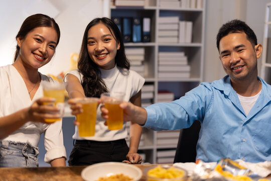 Friends toasting drinks celebrating party at office. Young Asian colleagues enjoying drinks and snacks together, celebrating a special occasion after work in the office.