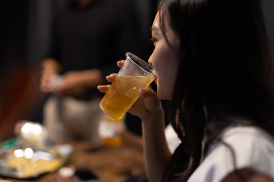 Young Asian woman enjoying a drink, having beer from a plastic cup at a casual social gathering indoors.