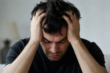 Fototapeta premium A man with dark, messy hair is holding his head in distress against a plain, light-colored background.