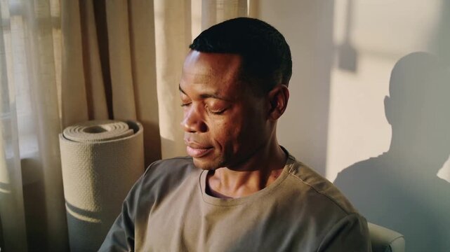 Sunlight falling causing man wearing earth tee turning head closing eyes near window, rolled mat