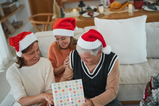 Senior couple opening Christmas gift together with adult daughter, smiling and sharing happy holiday moment during family gathering.