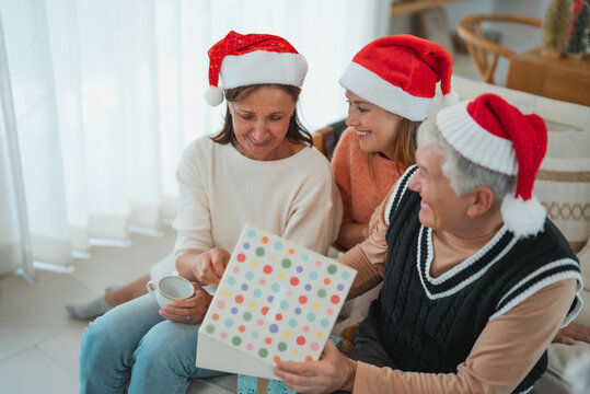 Senior woman opening Christmas present with husband and adult daughter, enjoying gift exchange and festive family time together at home.