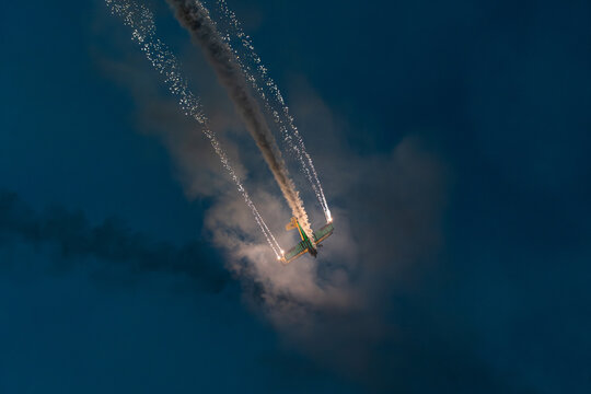 View of an aerobatic airplane performing at the Antidotum Airshow at Leszno Airport, leaving bright smoke trails and pyrotechnics in the night sky in Leszno, Wielkopolskie Voivodeship, Poland.