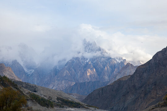 View of the Passu Cones mountain range partially obscured by clouds with rocky slopes and valley floor in Hunza Nagar, Gilgit, Pakistan.