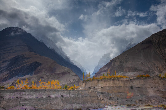 View of Sost village with dramatic mountain peaks, autumn trees with yellow foliage, and cloudy sky in Garelt, Gilgit, Pakistan.