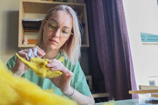 Young Woman With Glasses Carefully Shaping Bright Yellow Wool Into Small Felt Figure At Desk, Working In Cozy Room With Shelves, Curtain Light, And Soft Natural Daylight For Handmade Hobby