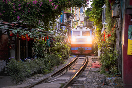 View of a train passing through the narrow Hanoi Train Street surrounded by residential buildings and lush hanging plants in Hoan Kiem, Ha Noi, Vietnam.