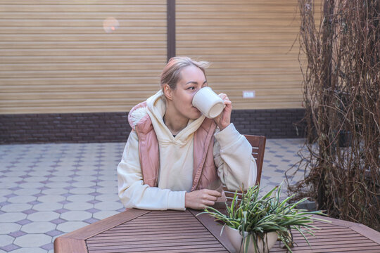 Young Woman In Hoodie And Pink Vest Drinks From White Mug While Sitting At Wooden Patio Table Beside Potted Plant, With Tiled Courtyard And Beige Wall Creating Calm Casual Outdoor,