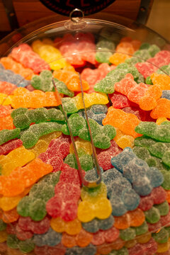 gummy bear candies are neatly arranged in a plastic bowl using metal tongs, creating a bright and playful display in an indoor setting, possibly at a candy store.