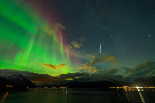 View of the Andoya Space rocket launch trail illuminated by vibrant green and purple northern lights over the dark waters and snow-capped mountains of Andenes, Nordland, Norway.