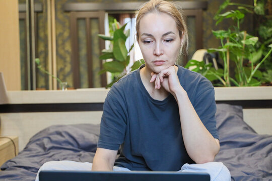 Young Woman In Casual Clothing Sits On Bed With Laptop, Chin Resting On Hand, Looking Pensive And Focused In Cozy Bedroom With Soft Bedding, Indoor Plants, And Warm Natural