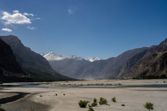 View of the majestic Haldi Cones with snow-capped peaks rising above a wide, dry riverbed under a clear blue sky in Khaplu, Gilgit, Pakistan.
