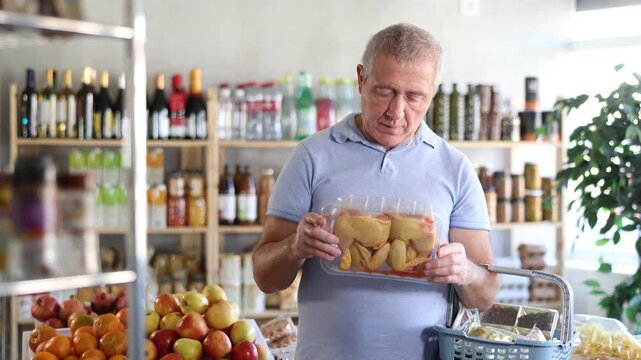 Senior man holding plastic containers with poultry half chicken carcass, thinking about buying. He choose ingredients for cooking dinner, thinking before buying, hesitating about choice chicken meat