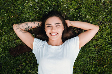 Fototapeta premium Smiling young woman with floral tattoo lying relaxed on green grass outdoors enjoying sunshine and carefree summer relaxation