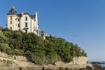 Fototapeta premium A grand Dinard house stands above the rocky shore, its steep roof and bright walls overlooking the calm sea under the clear blue sky.