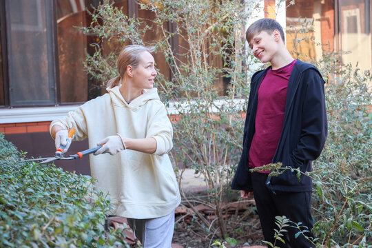 Two Young Adults Trim Overgrown Shrubs In Home Garden Beside House Windows, Smiling While Working Together With Long Handled Shears In Soft Daylight, Showing Outdoor Upkeep