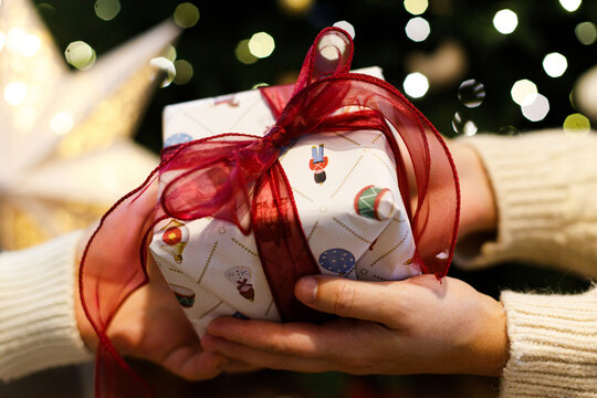 Two People Exchanging Wrapped Christmas Gift with Red Ribbon against xmas tree lights bokeh. Concept of Love and Holiday Present Giving. Happy Holidays