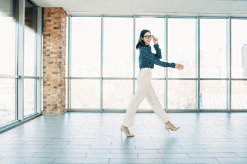 Fototapeta premium Confident businesswoman walking across a bright modern office space in heels and glasses, smiling as she strides with purpose toward a meeting or workspace