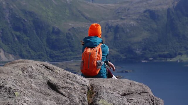 Aerial view of a female hiker wearing a backpack sitting on a rocky summit enjoying panoramic views of fjords and valleys in Lofoten, Norway.