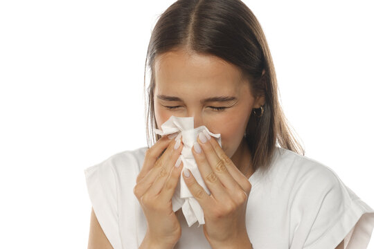 Portrait of a young woman sneezing into a tissue with closed eyes, on white background.