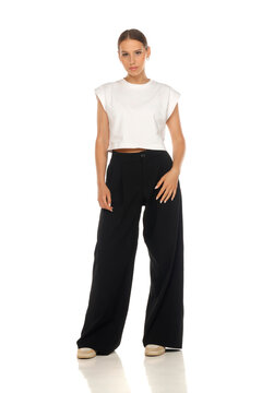 Full length fashion portrait of woman wearing white crop top and black trousers, standing pose, studio shot.