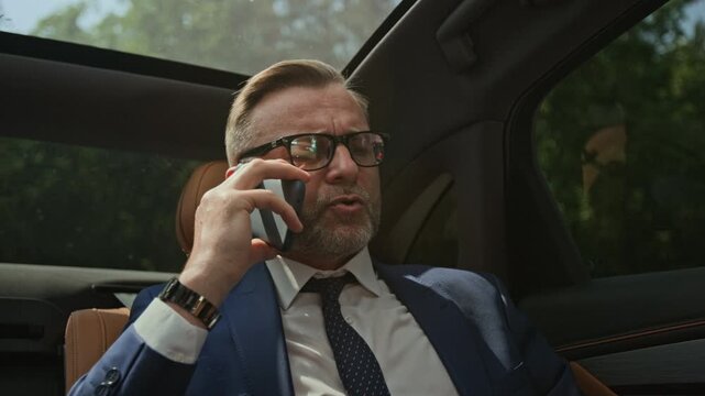 Middle aged Caucasian businessman talking on smartphone and gesturing while riding in backseat of luxury car. Clip is suitable for corporate communications and business travel promotions