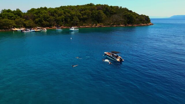 A picturesque scene unfolds in the adriatic sea, where a small boat glides near a lush, forested coastline. Several swimmers enjoy the crystal-clear waters, basking in the summer sun.