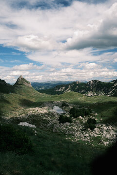 View of Durmitor National Park featuring rugged mountain peaks, lush green slopes, rocky terrain, and a small alpine pond under a cloudy sky in Montenegro.