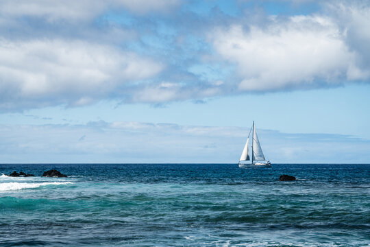 View of a sailboat sailing on the deep blue ocean with rocky outcrops and a cloudy sky in Las Galletas, Canary Islands, Spain.