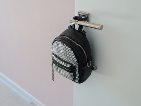 Small Black Backpack With Sparkling Rhinestone Front Panel Hanging From Sleek Metal Door Handle Beside Soft Pastel Wall And Stairway, Creating Stylish Minimalist Indoor Home Decor Scene With