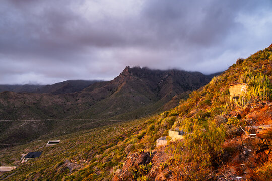 View of the rugged mountainous landscape of Costa Adeje at sunset, featuring rocky slopes covered in cacti, dramatic clouds, and small stone structures in the Canary Islands, Spain.