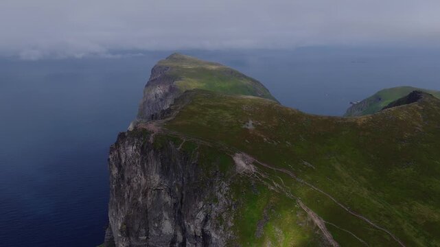 An aerial view captures the dramatic rocky sea cliffs covered in moss rising above the deep dark ocean water under an overcast sky in Lofoten, Norway.