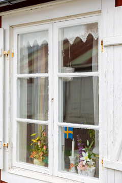 View of a traditional white wooden window with shutters, lace curtains, and colorful flowers in pots with a small Swedish flag in Gammelstaden, Norrbotten County, Sweden.
