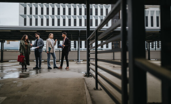 Four business colleagues stand outside a contemporary office talking and reviewing documents. The group discusses work together in an urban outdoor meeting scene.