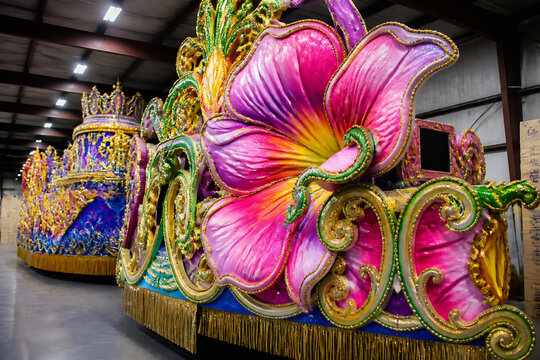 An elaborate, ornate float decorated with large pink and purple flowers is displayed indoors.