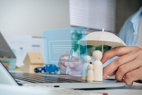 Hand protecting wooden family figures under umbrella while other hand signing digital contract document on laptop interface, concept of family insurance protection, life policy, and financial security