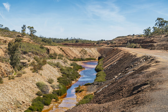 View of Minas de Sao Domingos, an abandoned mining site with a stream of reddish water flowing through rocky terrain under a clear blue sky in Corte do Pinto, Beja District, Portugal.