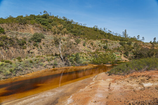 View of Minas de Sao Domingos with its characteristic orange-tinted stream flowing through the rugged landscape under a clear blue sky in Corte do Pinto, Beja District, Portugal.