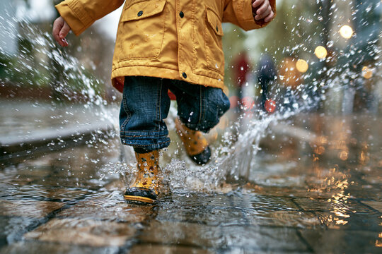 Low angle view of child in yellow raincoat and rubber boots splashing water in puddle on wet street