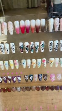 lateral camera pan across clear-rack salon display revealing rows of decorated artificial nail tips, movement is visible through shifting reflections and changing