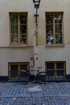 View of a black bicycle parked on a cobblestone street against a yellow building wall with windows and a vertical drainpipe in Stockholm, Stockholm County, Sweden.