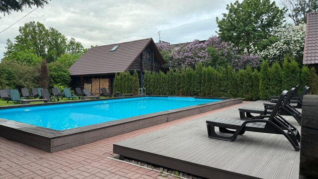 Outdoor rectangular swimming pool with bright blue water, surrounded by wooden decking, black sun loungers, green hedges, and a dark wooden cabin under a cloudy spring sky.