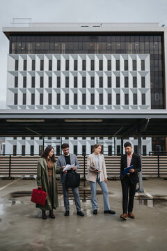 Four business colleagues stand together reviewing documents on a rooftop parking lot. A professional team consults papers with an office building facade in the background.