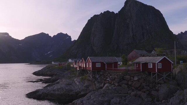 Traditional red wooden rorbuer cabins stand on stilts along the rocky coastline of A village in Lofoten, Norway, with steep mountains behind at sunrise.