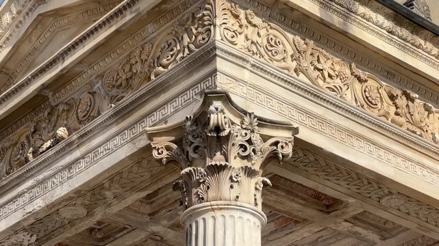 Close-up of a classical stone building facade featuring an ornate Corinthian column capital and intricately carved decorative frieze under bright sunlight.