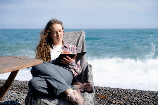 Young woman keeping a gratitude journal on a sunny seaside, writing in a notebook while enjoying a quiet moment outdoors. Concept of mindfulness, self-care, personal reflection and slow living.